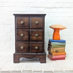 Puede incluir: Un mueble de madera marrón oscuro con seis cajones, cada uno con un pomo de latón. Una pila de libros coloridos y un objeto decorativo en forma de hongo naranja están junto al mueble. El mueble está sobre una superficie de ladrillo blanco.