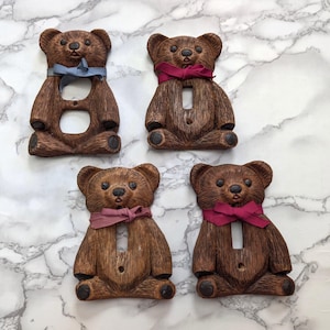 Pode incluir: Quatro espelhos de interruptor em forma de urso castanho com detalhes de pelo texturizado. Cada urso apresenta um pequeno laço de tecido atado ao pescoço em azul, rosa escuro ou malva. As placas possuem aberturas para interruptores ou tomadas.