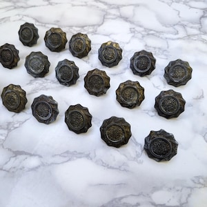 May include: A collection of sixteen circular metal buttons featuring a dark, weathered bronze finish. Each button has a scalloped, fluted edge and a textured, raised center with a detailed, intricate pattern visible on the surface of the metal.