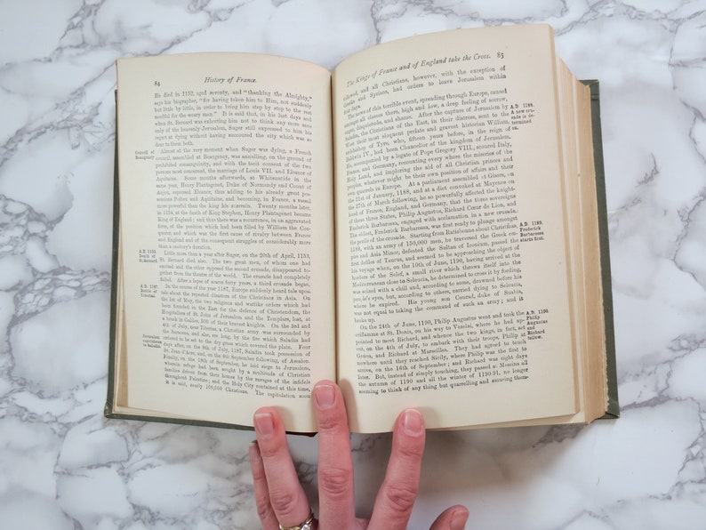 K&ouml;nnte beinhalten: Ein ge&ouml;ffnetes Buch mit gr&uuml;nem Einband. Die Seiten sind vergilbt und der Text ist schwarz. Das Buch tr&auml;gt den Titel "Geschichte Frankreichs" und "Die K&ouml;nige von Frankreich und England nehmen das Kreuz".