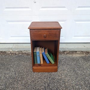 May include: A brown wooden nightstand featuring a single top drawer with a round knob and an open lower shelf containing five books with green, blue, and tan spines. The furniture piece has a rectangular top surface and a solid base construction.