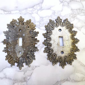 Peut inclure: Deux plaques d'interrupteur ovales en métal avec un motif de bordure en relief rappelant des feuilles. Les plaques présentent une finition métallique patinée, une découpe rectangulaire centrale et deux trous de vis circulaires.
