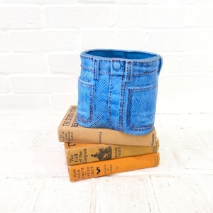 Op de afbeelding: Een blauwe keramische plantenbak in de vorm van een jeans, compleet met zakken, riemlussen en stikseldetails. De plantenbak staat op een stapel van drie vintage boeken met oranje en bruine covers.