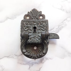 May include: An antique, ornate metal door latch with a keyhole and a decorative handle. The latch is dark gray with intricate floral and scrollwork details. The handle is a circular pull, and the latch has a small, curved lever.