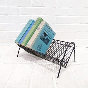 Könnte beinhalten: Ein schwarzer Bücherständer aus Metall mit mehreren alten Büchern. Die Bücher haben blaue, grüne und weiße Einbände. Der Bücherständer hat ein Rautenmuster und ist abgewinkelt, mit vier Beinen. Der Hintergrund ist eine weiße Backsteinmauer.