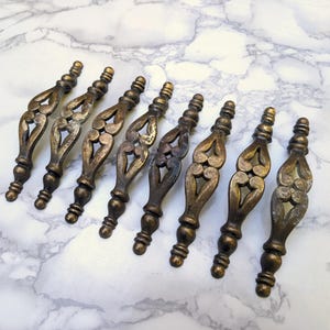May include: A set of seven elongated metal drawer pulls with a dark, weathered bronze finish. Each handle features a central openwork design with a symmetrical, heart-like cutout pattern, flanked by rounded, tiered finials at both ends.