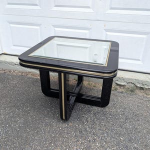 May include: A square end table with a glossy black finish and a glass top insert. The table features rounded corners and a gold-toned metal trim accent that runs along the edge of the tabletop and down the center of the intersecting base legs.