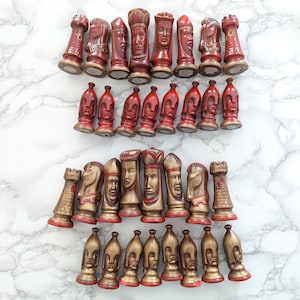 May include: A collection of vintage chess pieces in red and gold. The pieces are arranged on a white marble surface. The chess pieces include rooks, knights, bishops, pawns, and a king and queen. The pieces have detailed facial features.