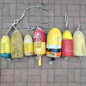May include: A collection of seven weathered cylindrical fishing buoys in various sizes and colors, including yellow, red, and white. Several buoys feature visible identification numbers like 1064, 4052, and 73, with attached ropes and metal hardware.