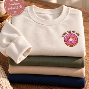 Könnte beinhalten: Ein Stapel aus vier gefalteten Sweatshirts mit Rundhalsausschnitt in Creme, Olivgrün, Beige und Marineblau. Das oberste cremefarbene Sweatshirt zeigt einen gestickten rosa Donut mit bunten Streuseln und den Text Donut Kill My Vibe in Braun.