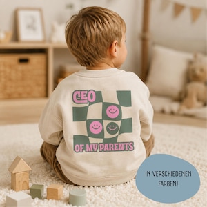 Könnte beinhalten: Ein beigefarbenes Langarm-Sweatshirt mit einem Grafikdruck auf der Rückseite, bestehend aus einem grün-beigen Karomuster, rosa Smileys und dem Schriftzug "CEO OF MY PARENTS" in rosa Buchstaben. Das Sweatshirt hat einen Rundhalsausschnitt.