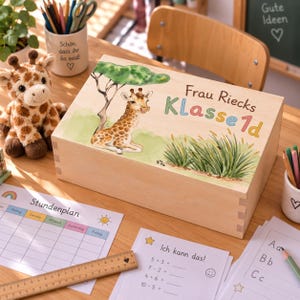 Könnte beinhalten: Eine rechteckige Holzkiste mit einem aufgedruckten Bild einer Giraffe, die unter einem Baum im hohen Gras sitzt. Der Text "Frau Riecks Klasse 1d" ist in bunten Buchstaben auf den Deckel gedruckt. Die Kiste hat sichtbare Fingerzinken an den Ecken.