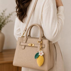 May include: A beige structured handbag with a textured finish, featuring a gold-tone metal turn-lock closure and a detachable shoulder strap. A small, crocheted lemon charm with a white flower and green leaf is attached to the handle hardware.