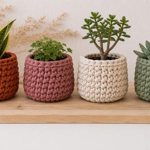 Puede incluir: Cuatro macetas de ganchillo en tonos naranja, malva, crema y verde salvia, cada una con una planta diferente. Las macetas están dispuestas sobre una superficie de madera, mostrando una variedad de texturas y colores.