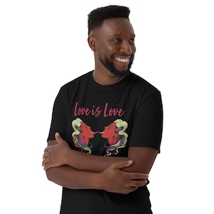 May include: Black t-shirt with the text "Love is Love" in pink script. The shirt features a graphic of two red faces with tongues touching, with rainbow-colored hair. The person wearing the shirt is smiling.