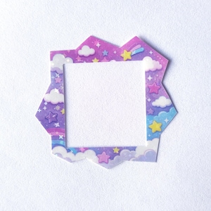 Può includere: Una cornice di forma quadrata con un bordo esterno frastagliato a forma di stella, caratterizzata da uno sfondo con sfumature viola e blu. Il design include nuvole bianche, varie stelle gialle, rosa e blu e piccoli accenti scintillanti.