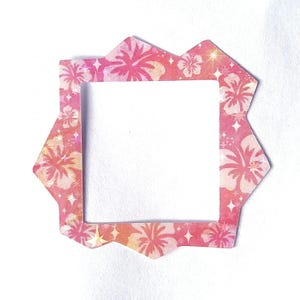 May include: A square-shaped frame with an irregular, jagged outer edge. The frame features a vibrant pink and orange background decorated with a repeating pattern of hibiscus flowers and small, four-pointed star shapes scattered throughout.