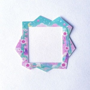 Puede incluir: Un marco de forma cuadrada con un borde exterior irregular y dentado que presenta un diseño en tonos pastel de nubes rosas, tallos de bambú verdes, flores pequeñas, una luna creciente, estrellas, un puente y un pequeño edificio sobre un fondo verde azulado.