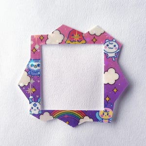 Puede incluir: Un marco decorativo de múltiples lados con un diseño de gradiente púrpura y rosa. El marco presenta personajes de dibujos animados, nubes, estrellas y un arcoíris. El marco está hecho de un material brillante y holográfico.