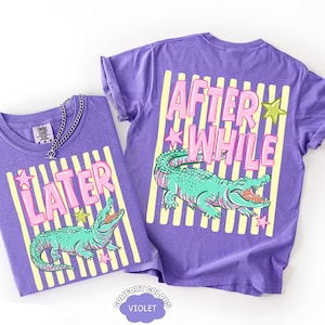 May include: Two violet t-shirts with a graphic design. One shirt reads "LATER" and the other reads "AFTER WHILE" in pink letters. Both shirts feature a teal alligator illustration and yellow stripes.