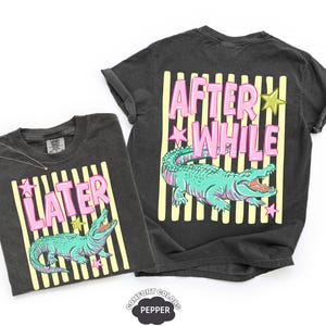 May include: Two dark gray t-shirts with a graphic of a turquoise alligator and the text "LATER" and "AFTER WHILE" in pink letters. The shirts have yellow vertical stripes and star accents.