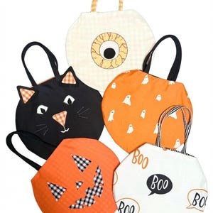 May include: Five round fabric tote bags with Halloween themes. One features a large eyeball, one a black cat face, one an orange ghost pattern, one a jack-o'-lantern face with gingham details, and one a white background with the word BOO in speech bubbles.