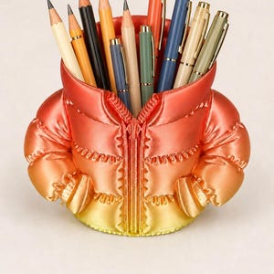 Op de afbeelding: Een bureau-organizer in de vorm van een pufferjack met een kleurverloop van oranje aan de bovenkant naar geel aan de onderkant. Het jack heeft een centrale ritssluiting en twee zijzakken, gevuld met diverse potloden en pennen.
