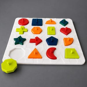 Op de afbeelding: Een wit vierkant sorteerbord met een raster van uitsparingen. Het bord bevat diverse kleurrijke geometrische stukken, waaronder een rode cirkel, blauw vierkant, oranje halve cirkel, groene ruit, plusteken, zeshoek, rechthoek, hart, ster, pijl, vijfhoek en halve maan.
