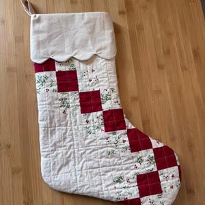 Könnte beinhalten: Ein gesteppter Weihnachtsstrumpf mit einem diagonalen Schachbrettmuster aus einfarbigen roten Quadraten und weißen Stoffquadraten, die mit grünen Stechpalmenblättern und roten Beeren bedruckt sind. Der Strumpf hat einen cremefarbenen Umschlag.
