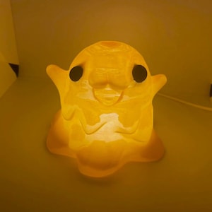 May include: A translucent, amber-coloured, glowing lamp shaped like a small, rounded creature with two dark, circular eyes. The figure has a smooth, moulded surface with subtle facial features and small, outstretched arms, emitting a warm light.