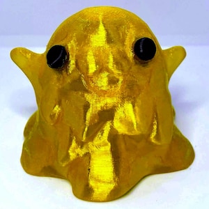 Puede incluir: Una pequeña figura tridimensional con forma de gota o fantasma, con un acabado dorado metálico. El objeto presenta una parte superior lisa y redondeada, dos pequeñas protuberancias laterales puntiagudas y dos ojos negros circulares.