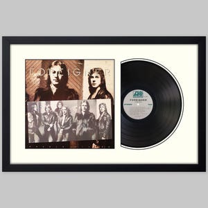 May include: A framed display featuring the Foreigner album cover art alongside a black vinyl record. The album cover shows the band members in a sepia-toned photograph with the text Foreigner. The vinyl record has a central Atlantic label.