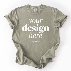 May include: A sage green short-sleeve t-shirt with a crew neckline. The center of the shirt features the white text "your design here" in a serif font, with the smaller white text "MagicMocks" printed directly below the main phrase.