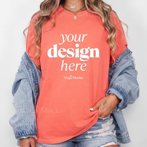 May include: A coral-colored short-sleeve t-shirt featuring the white text "your design here" and "MagicMocks" printed on the front. The shirt is worn under a light blue denim jacket with distressed denim shorts visible at the bottom hem.