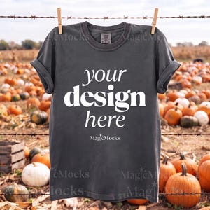 Może przedstawiać: Ciemnoszary t-shirt z krótkim rękawem zawieszony na drewnianym klamerce na drucie. Koszulka posiada biały napis "YOUR design here" wykonany pogrubioną czcionką szeryfową, a poniżej znajduje się mniejszy biały napis "MagicMocks".