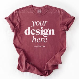 May include: A short-sleeved crew neck t-shirt in a muted dark red color. The front of the shirt features the white text "your design here" in a bold serif font, with the smaller white text "MagicMocks" printed directly below the main phrase.