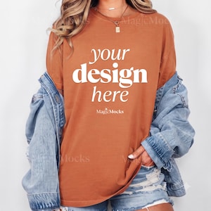 May include: A burnt orange short-sleeved t-shirt featuring the white text "your design here" and "MagicMocks" printed on the front. The t-shirt is worn with a light blue denim jacket and light blue denim shorts, creating a casual layered look.