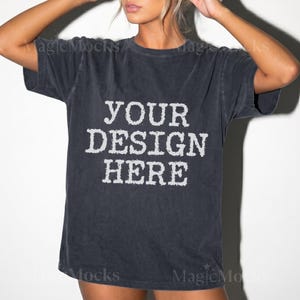 Może przedstawiać: Koszulka z krótkim rękawem w kolorze ciemnoszarym z wykończeniem w stylu vintage. Na środku koszulki znajduje się biały napis "YOUR DESIGN HERE" wykonany pogrubioną, postarzaną czcionką blokową. Odzież posiada okrągły dekolt i luźny krój.