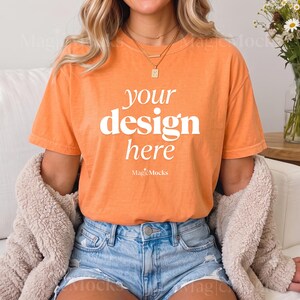 May include: An orange short-sleeve t-shirt featuring the white text "your design here" and "MagicMocks" printed on the front. The shirt is worn with light blue denim shorts and a beige fuzzy jacket, paired with a gold necklace with a letter K pendant.
