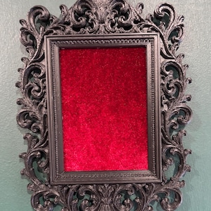 Puede incluir: Un marco de fotos rectangular con un borde negro ornamentado de plástico con intrincados adornos y tallas florales. El centro del marco está relleno con una inserción de tela de terciopelo rojo vibrante, creando un contraste con el marco.