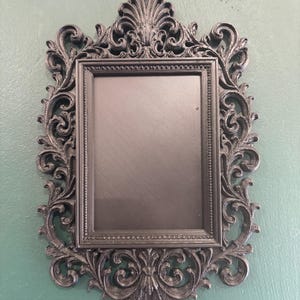 Puede incluir: Un marco de fotos rectangular negro con un borde ornamentado de volutas intrincadas y motivos detallados de hojas de acanto en la parte superior e inferior. El marco tiene un borde interior texturizado y un panel central liso y plano, de aspecto oscuro.