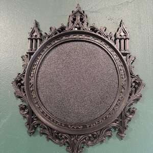 May include: A circular black cork board framed in an ornate, dark matte plastic structure featuring intricate architectural details, including pointed arches, spire-like towers, and elaborate scrollwork along the outer decorative border.