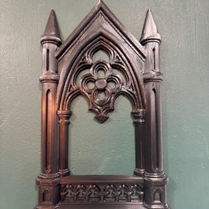 May include: A dark brown, gothic-style decorative frame. The frame features intricate architectural details, including pointed arches, spires, and a quatrefoil design. The frame is made of a solid material and is designed to be mounted on a wall.
