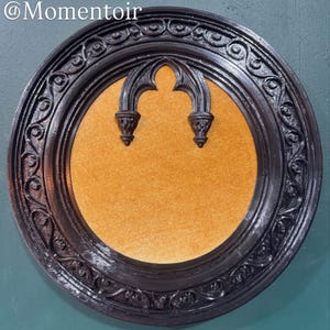 May include: A round black frame with intricate scrollwork relief patterns surrounding a central orange cork board surface. The frame features a decorative black arch-shaped element at the top center. The text @Momentoir is visible in the top left.