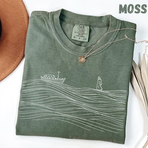 Könnte beinhalten: Moosgrünes T-Shirt mit einer weißen Strichzeichnung eines Bootes auf welligem Wasser und einer Figur, die geht. Das Shirt hat einen Rundhalsausschnitt und ein kleines Etikett. Eine goldene Halskette mit Anhänger befindet sich auf dem Shirt.