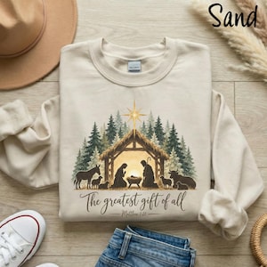 May include: A sand-coloured crewneck sweatshirt featuring a printed nativity scene with a stable, figures, and animals under a star. The text reads The greatest gift of all and Matthew 1:21. The design is set against a background of evergreen trees.