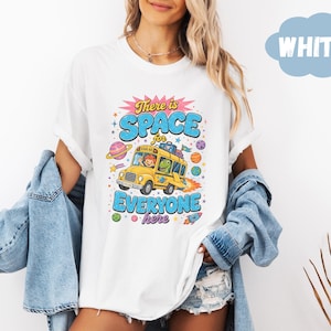 Op de afbeelding: Een wit t-shirt met een afbeelding van een gele schoolbus met een kind en een dinosaurus als passagiers. De tekst luidt There is SPACE for EVERYONE here. Het ontwerp bevat kleurrijke planeten, sterren en een raket op een witte achtergrond.
