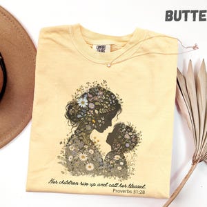 Puede incluir: Camiseta Comfort Colors de color mantequilla con una silueta floral de madre e hijo. El diseño incluye el texto "Her children rise up and call her blessed. Proverbs 31:28". También se ve un collar de oro con un colgante de estrella.
