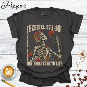 Peut inclure: Un t-shirt gris anthracite avec une impression graphique représentant un squelette entouré de fleurs colorées. Le texte EZEKIEL 37:1-10 figure en haut et le texte DRY BONES COME TO LIFE est imprimé en bas du motif rectangulaire.