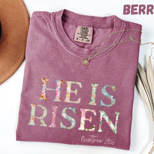 Könnte beinhalten: Beerenfarbenes T-Shirt mit dem Schriftzug "HE IS RISEN" in floralem Design. Der Text "Matthew 28:6" steht darunter. Eine goldene Halskette liegt über dem Shirt.